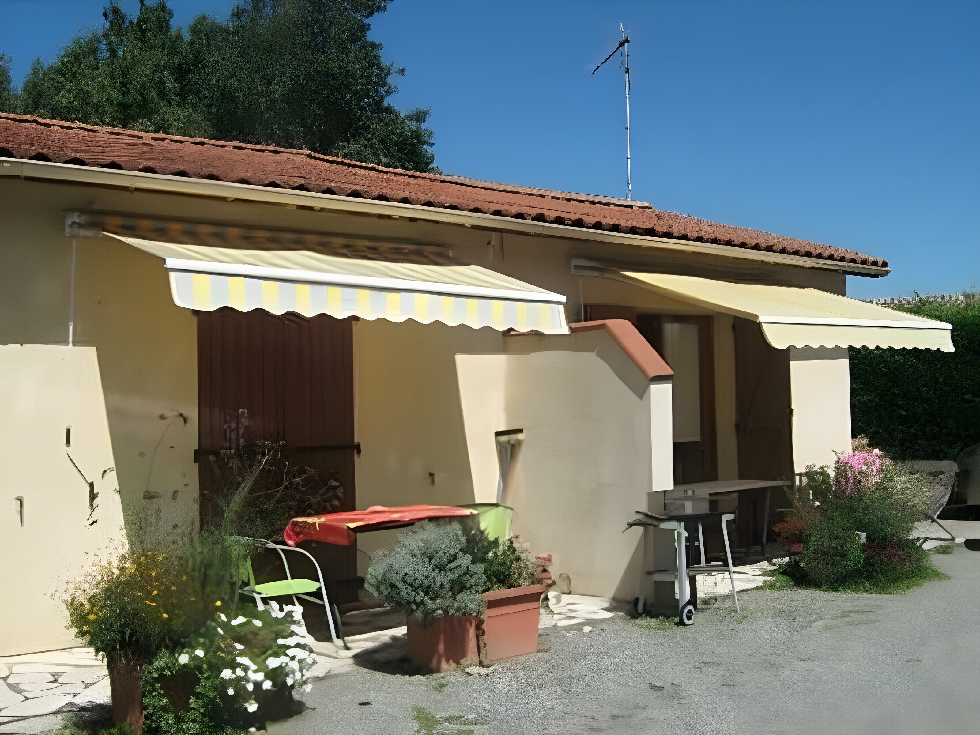 Une maison avec un toit recouvert de tuiles rouges, un petit porche doté d’un auvent jaune, une girouette et une allée gravillonnée.