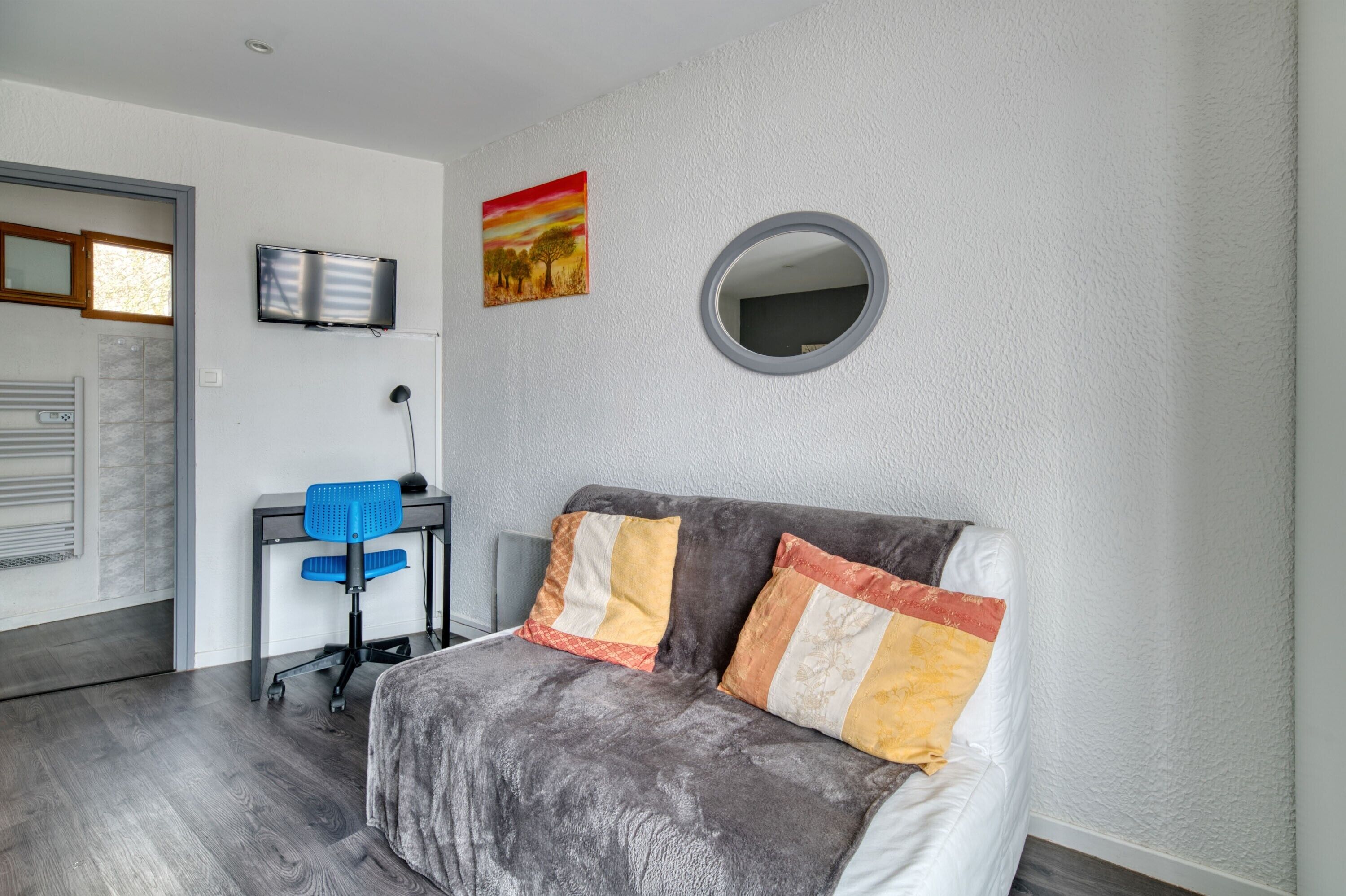 Ein modernes Zimmer mit grauem Sofa, Schreibtisch mit Stuhl, Flachbildfernseher, rundem Spiegel und einem Gemälde an der Wand.