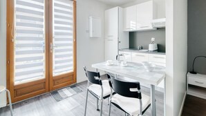 A modern kitchen with white cabinets, a dining table with chairs, and a large window with blinds. - Studio Apartment 'Le Laurier' with Private Terrace, Shared Garden and Wi-Fi (Lherm)