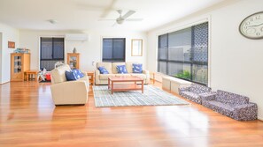 A spacious living room with a sofa, coffee table, and a patterned ottoman. There is a ceiling fan, a large window with blinds, and a clock on the wall.