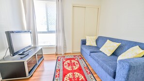 A living room with a TV, a blue sofa with yellow pillows, a red patterned rug, and a window with blinds.