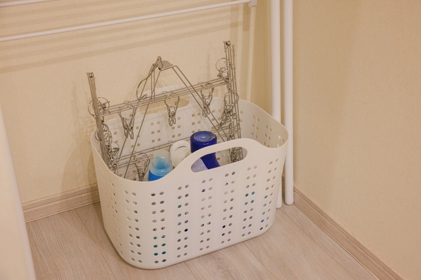 Una cesta de ropa blanca con perchas metálicas y una botella azul, ubicada frente a una pared con una tubería blanca.