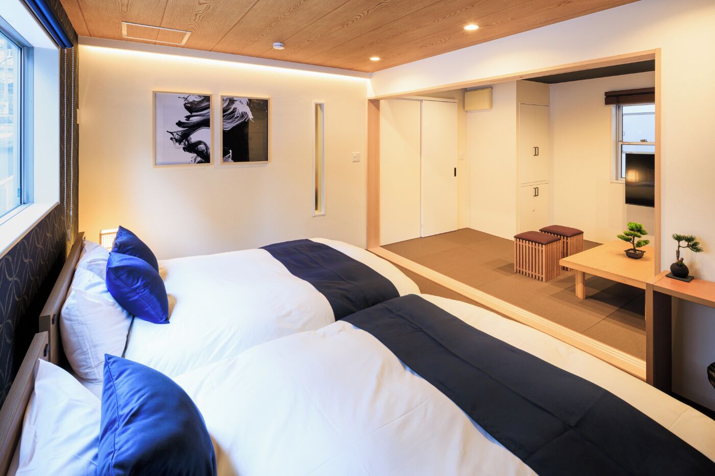 Ein modernes Hotelzimmer mit zwei Betten, einem kleinen Tisch mit einer Bonsai-Pflanze und Blick auf die Stadt durch das Fenster.