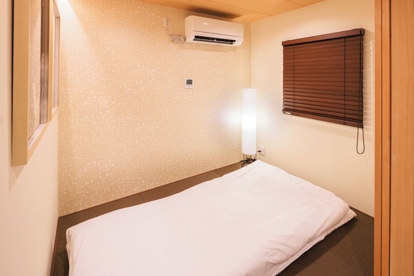 A small, neatly arranged room with a bed, an air conditioning unit, a wall-mounted light, and a window with blinds.