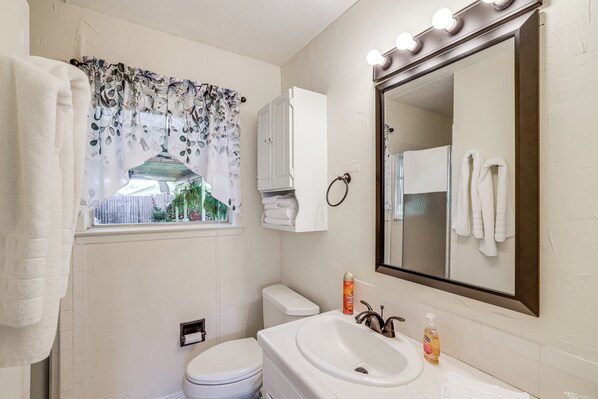 A bathroom with a toilet, sink, and mirror. There is a window with a curtain, and a towel rack with towels.