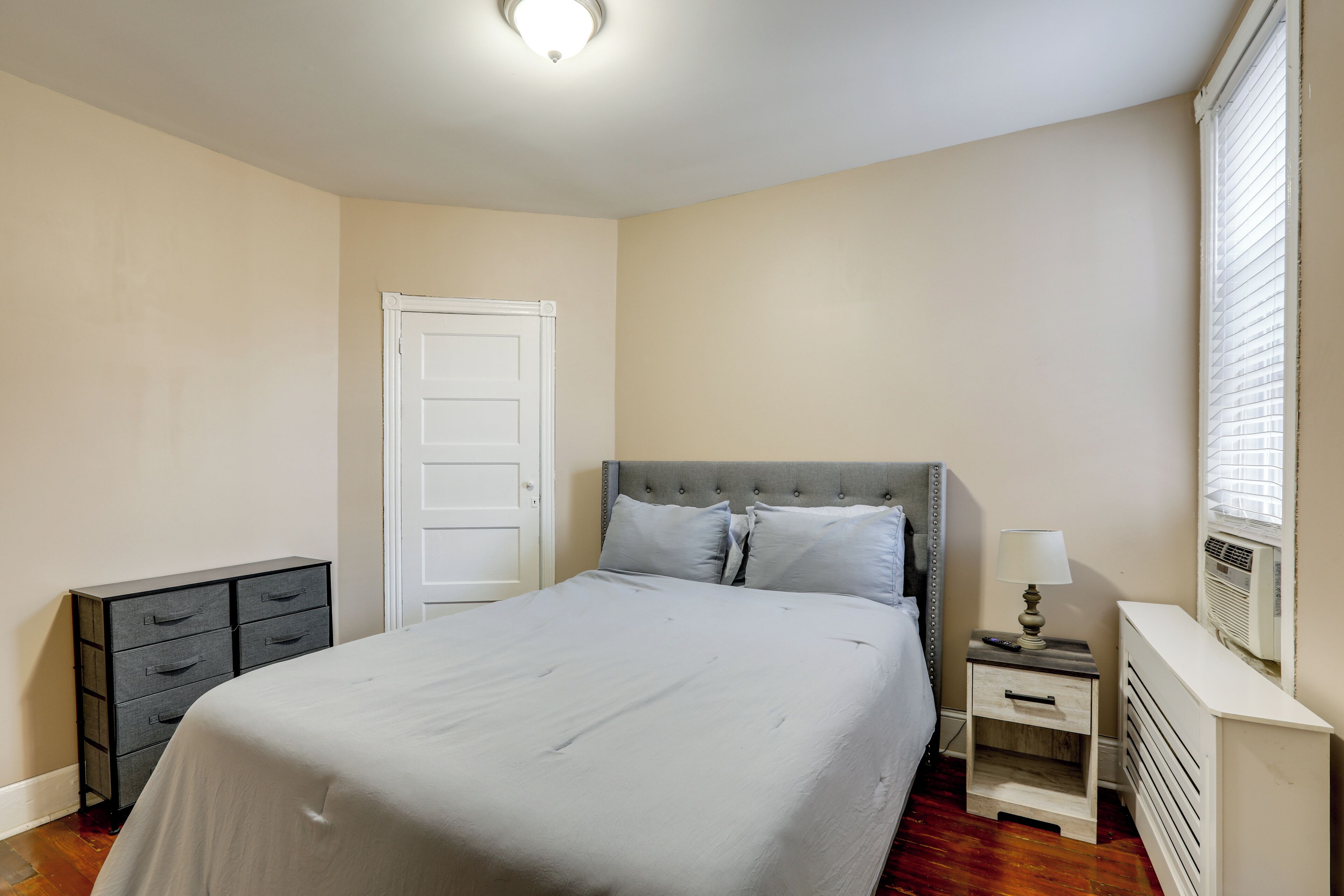 Un dormitorio con cama, cómoda, mesita de noche con lámpara, puerta y radiador.