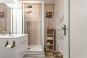 Ein Badezimmer mit weißem Waschbecken, Spiegel und Holzschrank. Es gibt eine Dusche mit Holztür und einen Handtuchhalter mit Handtüchern.