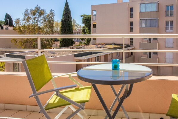 Ein Balkon mit einem grünen Stuhl, einem kleinen runden Tisch und einem blauen Gegenstand, mit Blick auf Gebäude und Bäume.