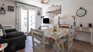 A living room with a sofa, a dining table with chairs, a TV, a lamp, and a painting on the wall. - Apartment "400m from the Beach" with Balcony (Fréjus)