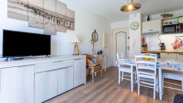 A modern kitchen with a flat-screen TV, wooden cabinets, a dining table with chairs, and a hanging light fixture. - Apartment "400m from the Beach" with Balcony (Fréjus)