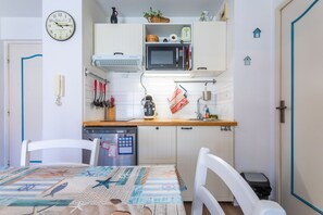 Cozinha compacta com armários brancos, micro-ondas e pia. Há um relógio na parede e uma mesa com toalha de mesa estampada.