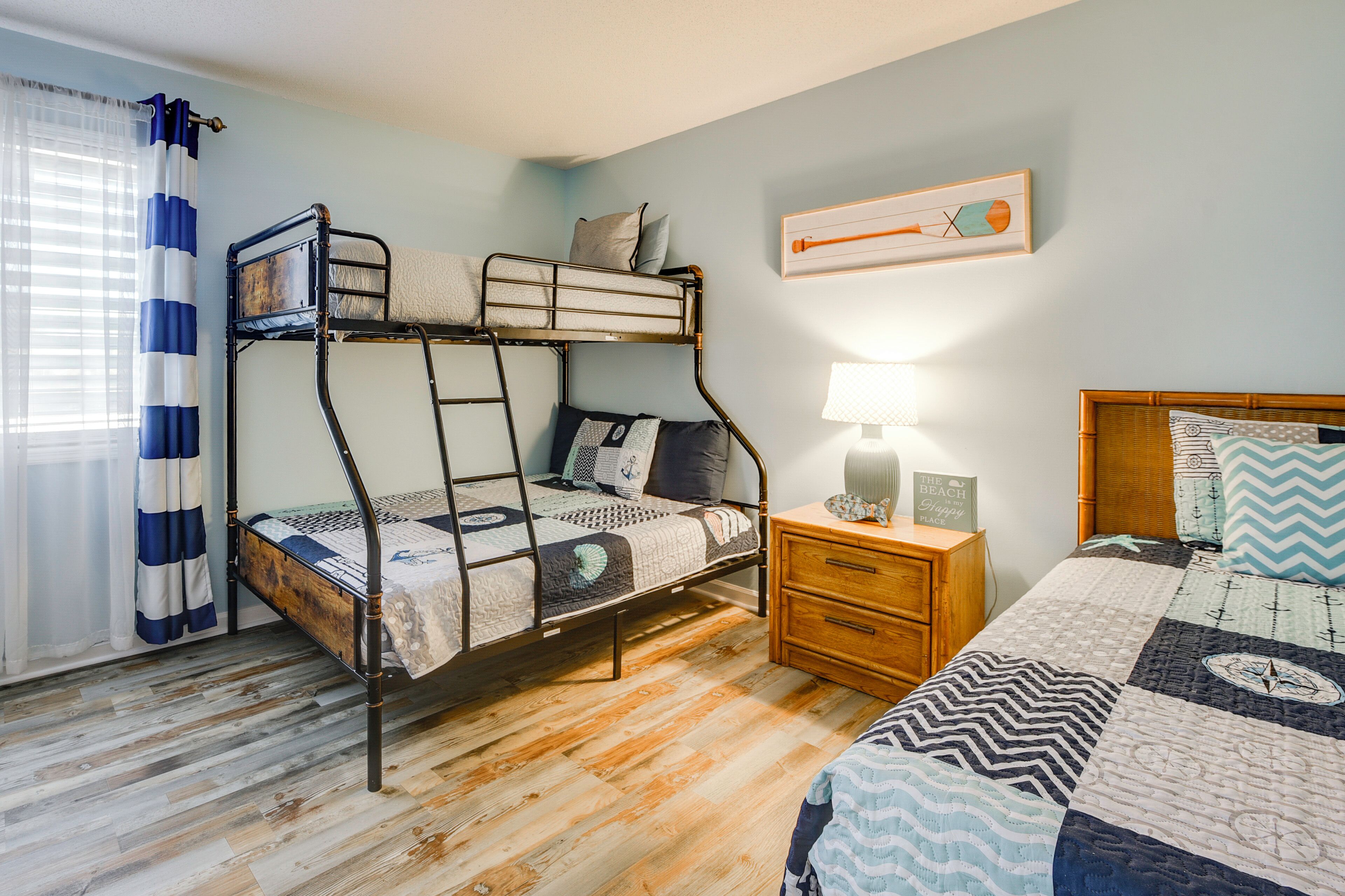 Un lit superposé avec une structure en bois, une table de chevet avec une lampe, une commode en bois et un tableau accroché au mur.