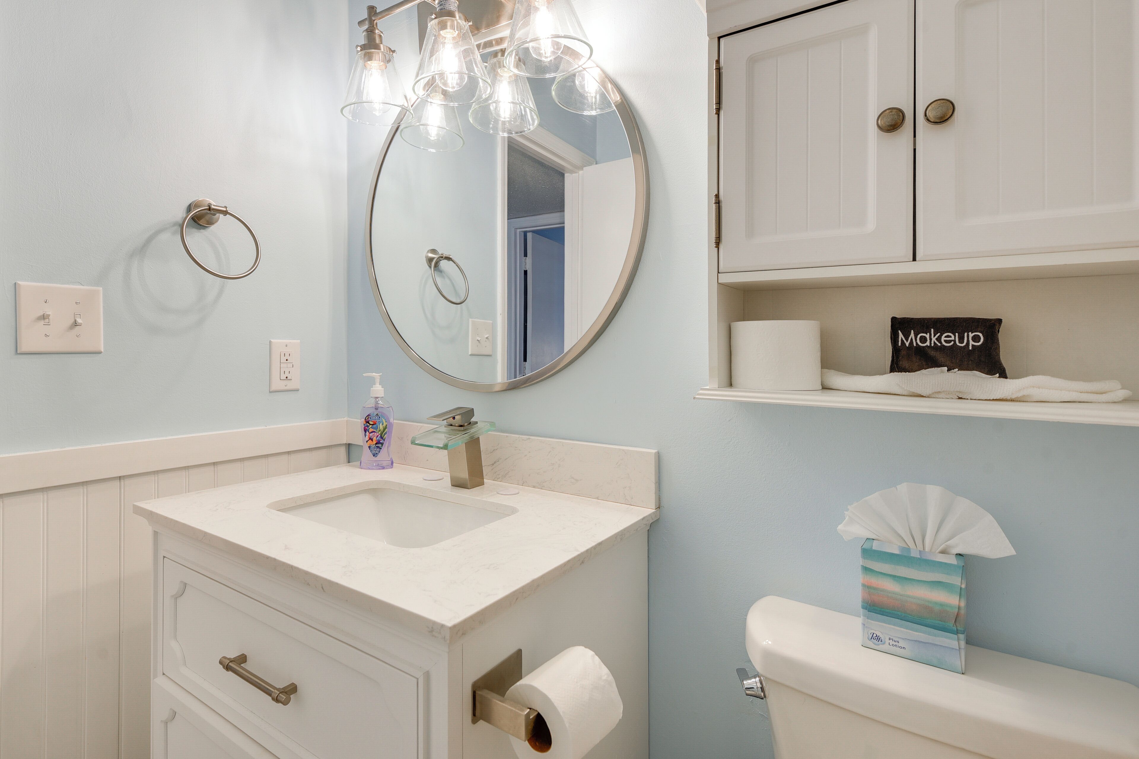 A bathroom with a white sink, a round mirror, a toilet, and a shelf with a makeup box.