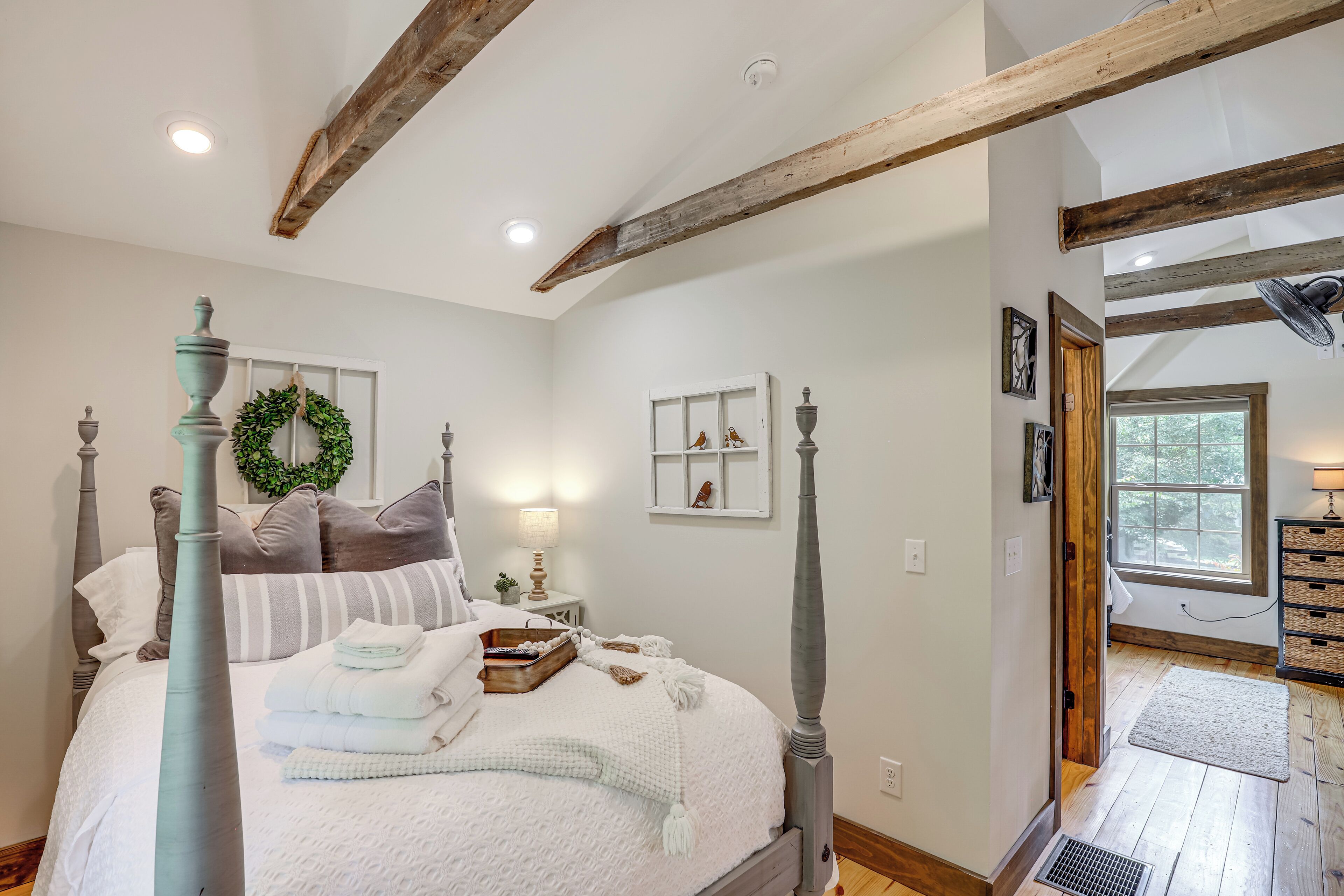 A bedroom with a four-poster bed, a wooden ceiling, a window with a view, and a small bedside table with a lamp.