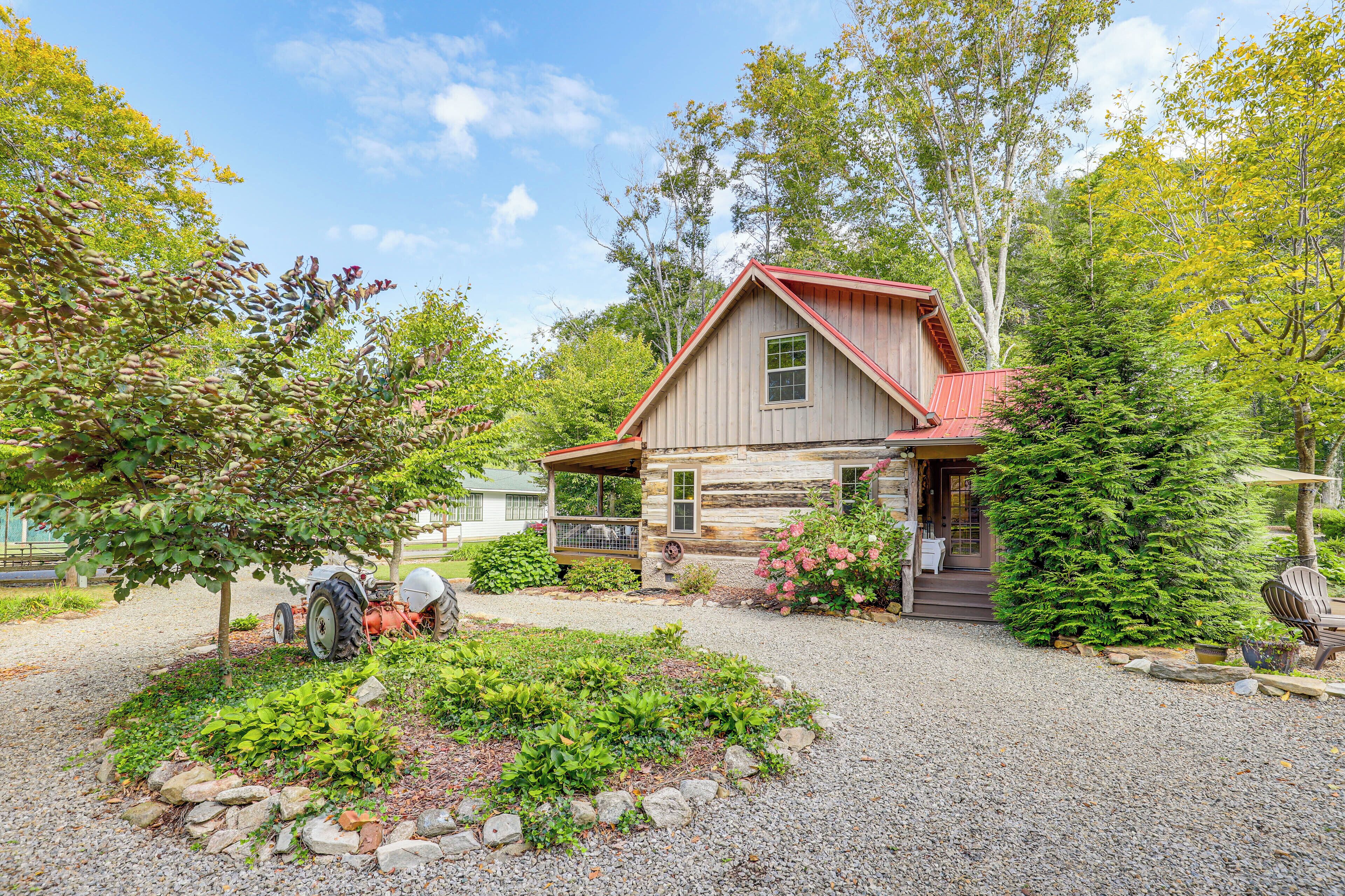 A rustic log cabin with a red roof, surrounded by a gravel driveway and lush greenery.