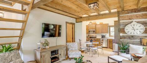 A spacious living area with a high ceiling, wooden beams, a television mounted on the wall, and a kitchen in the background.