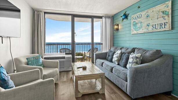 Una sala de estar con un sofá gris, dos sillones, una mesa de centro de madera y un ventanal con vistas al mar.