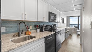 Una cocina moderna con armarios blancos, un lavamanos de acero inoxidable y un microondas. Se ve un comedor con una mesa y sillas al fondo.