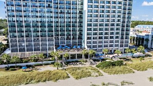 Un hotel de gran altura con fachada de vidrio moderna, un área de piscina con sombrillas y una playa en primer plano.