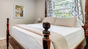 A bedroom with a large bed, wooden headboard, a window with patterned curtains, and a wall-mounted abstract painting.
