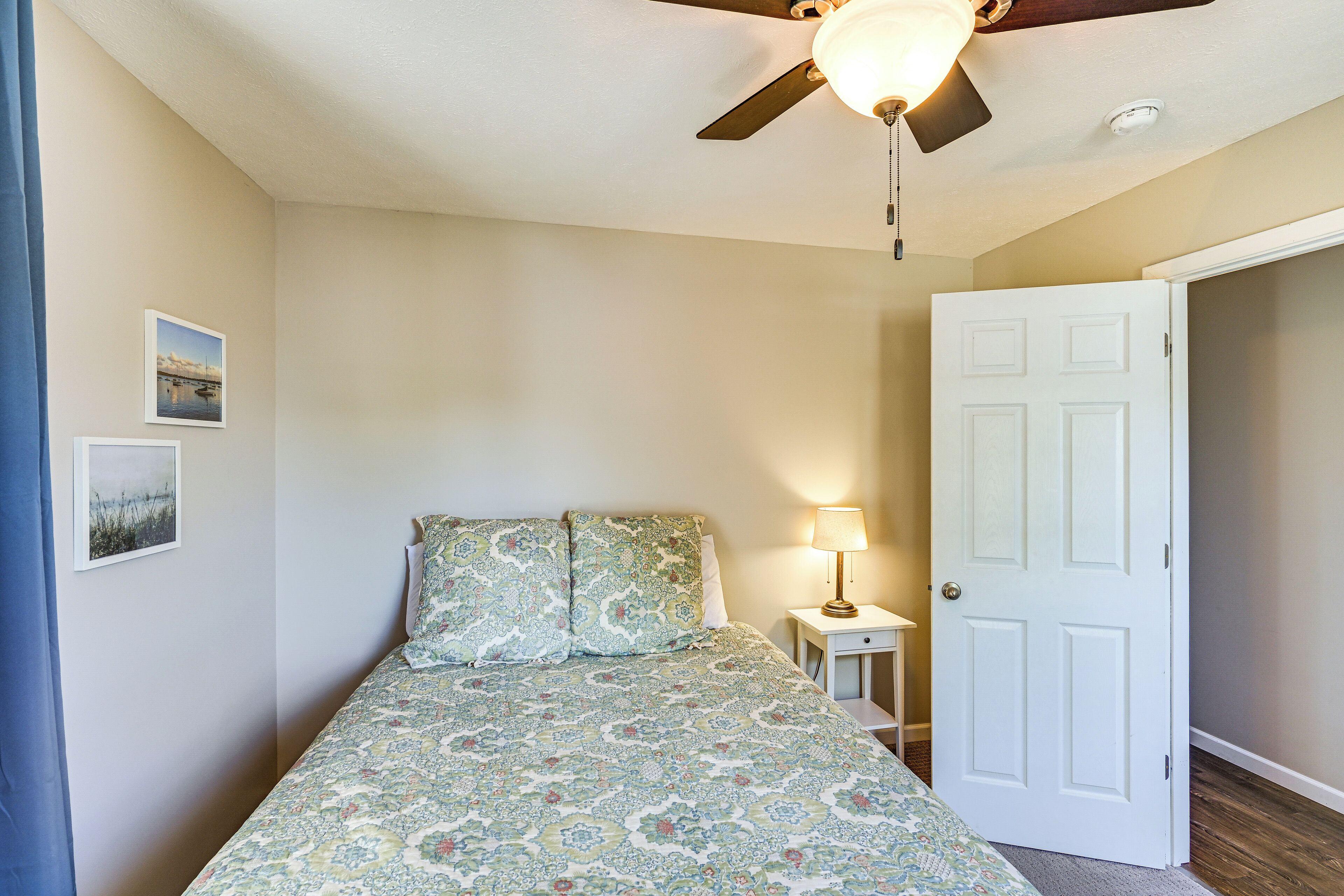 A bedroom with a bed, a ceiling fan, a nightstand with a lamp, and a door.