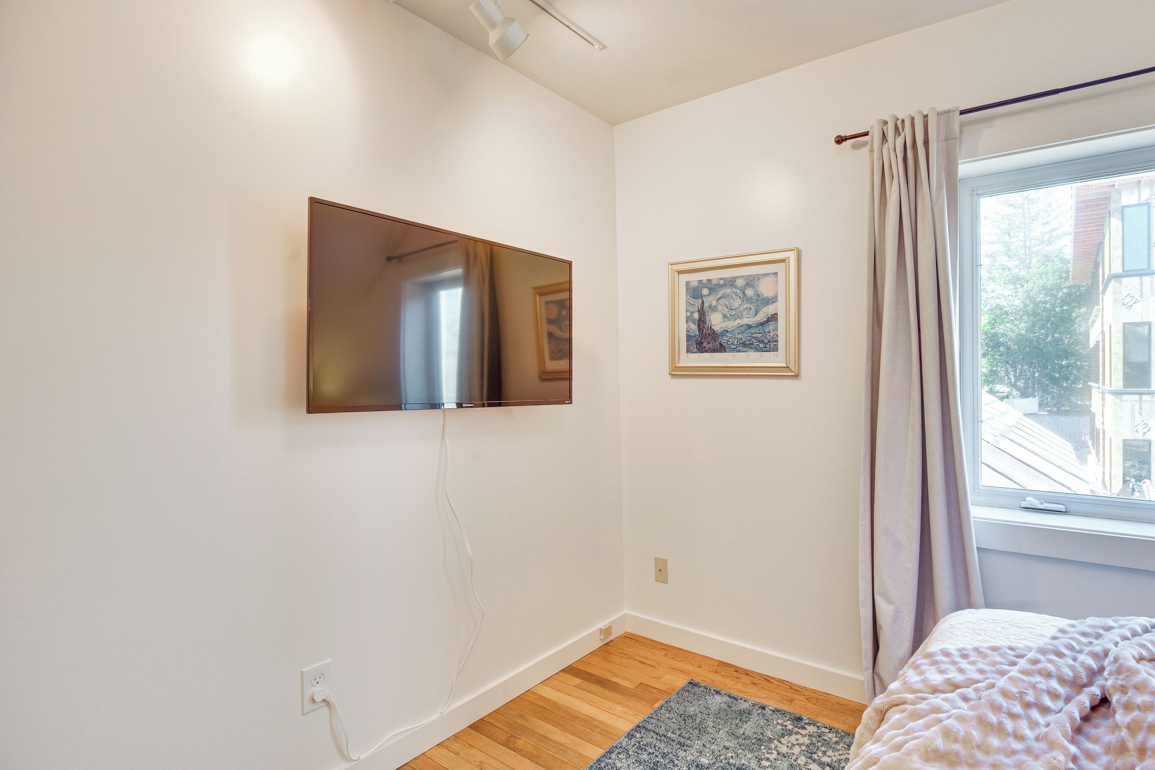 A bedroom with a bed, a window with curtains, a mounted TV, and a framed picture on the wall.
