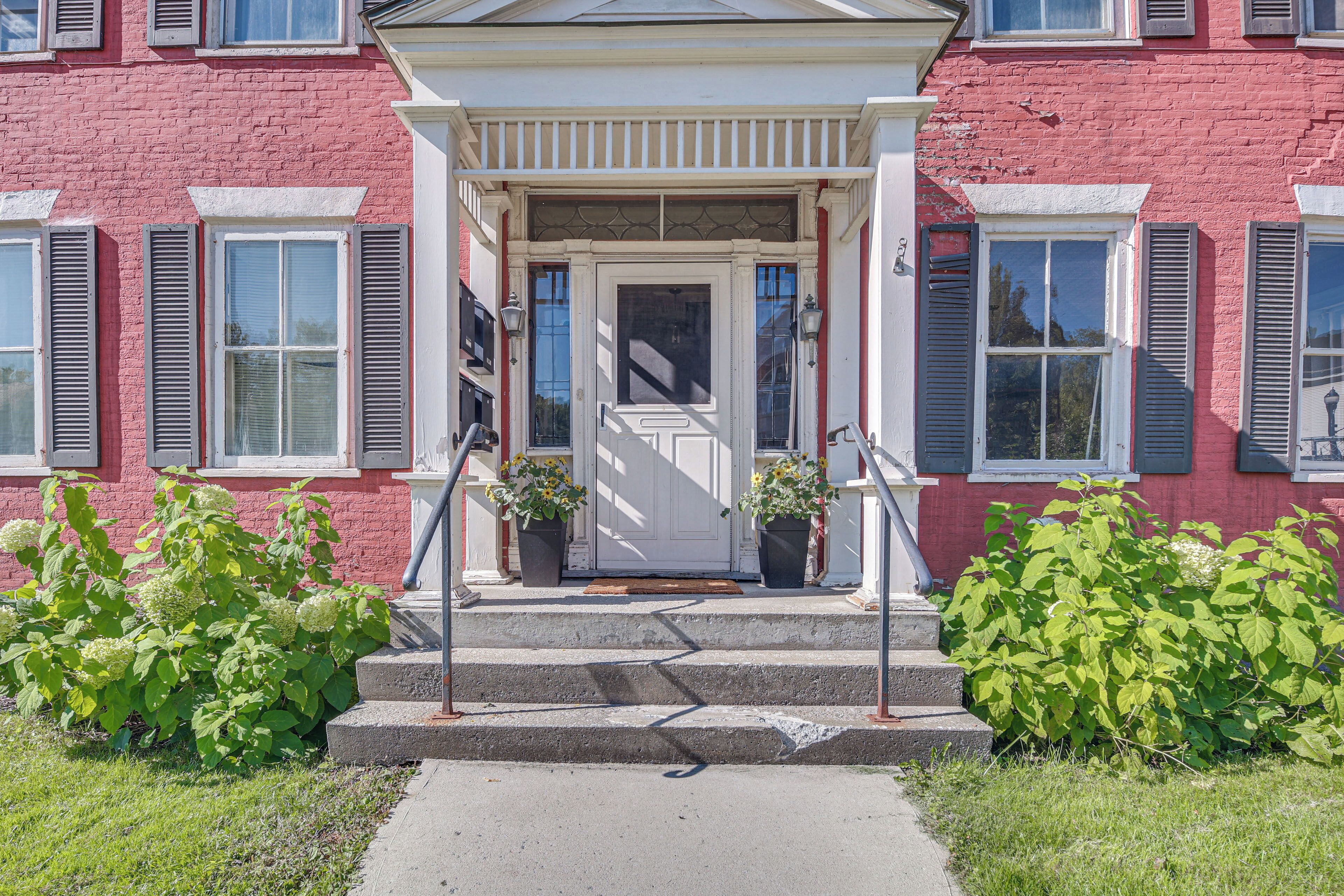 A red brick building with a white door, two potted plants, and a set of concrete steps leading to the entrance.