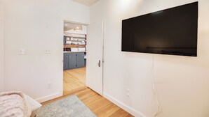 Un appartement moderne doté d’un grand téléviseur à écran plat fixé au mur, d’un coin cuisine à l’arrière-plan et d’un plancher en bois.