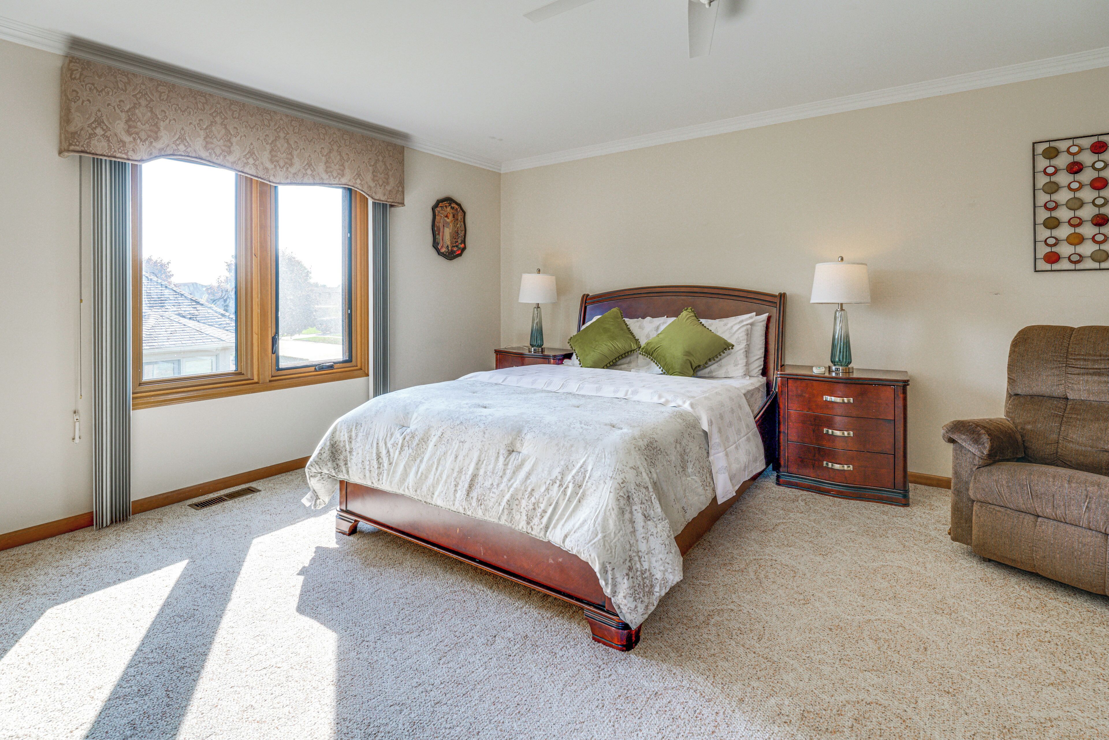 A bedroom with a large bed, two nightstands, a dresser, a chair, a window with curtains, and a wall decoration.