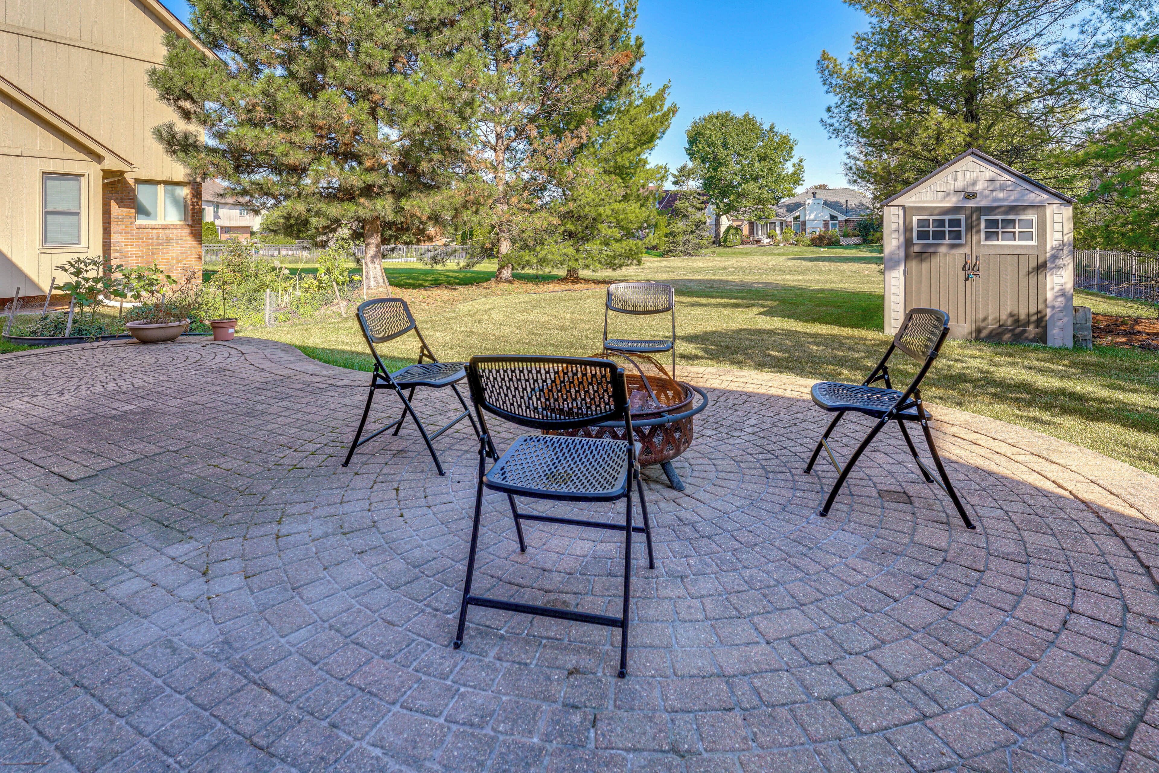 A patio with four metal chairs and a small table, surrounded by a brick patio and a lawn with trees.