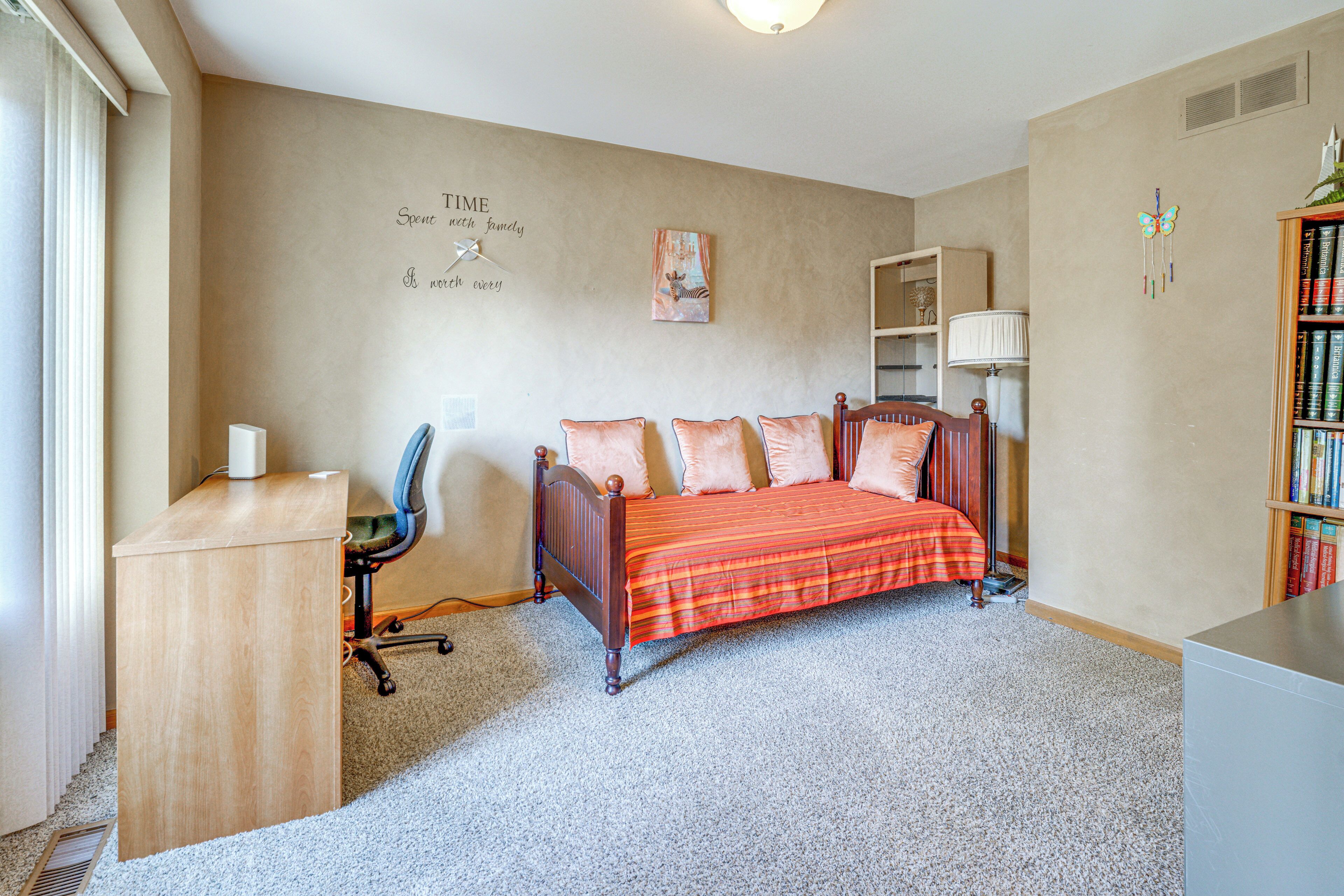 A bedroom with a bed, a desk, a chair, a bookshelf, and a wall with a decorative piece.