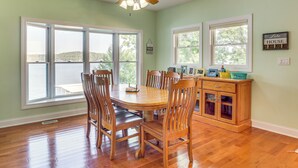 A dining area with a wooden table and chairs, large windows with a view of the water, and a wooden cabinet with kitchen items. - Water-view Deck! Family Home on Lake of the Ozarks (Roach)