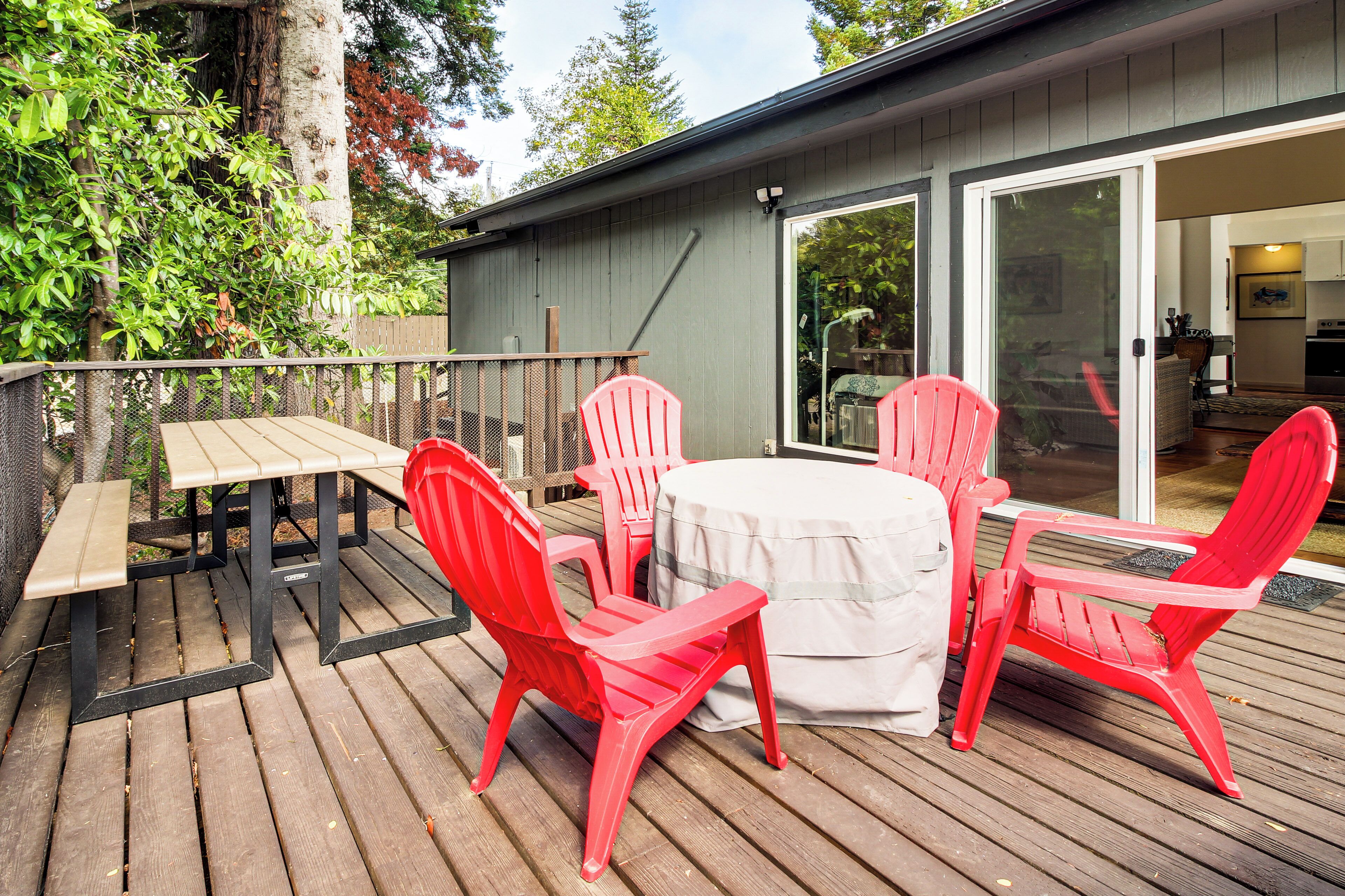 A wooden deck with a table and four red chairs, a sliding glass door leading to a kitchen, and a tree in the background.
