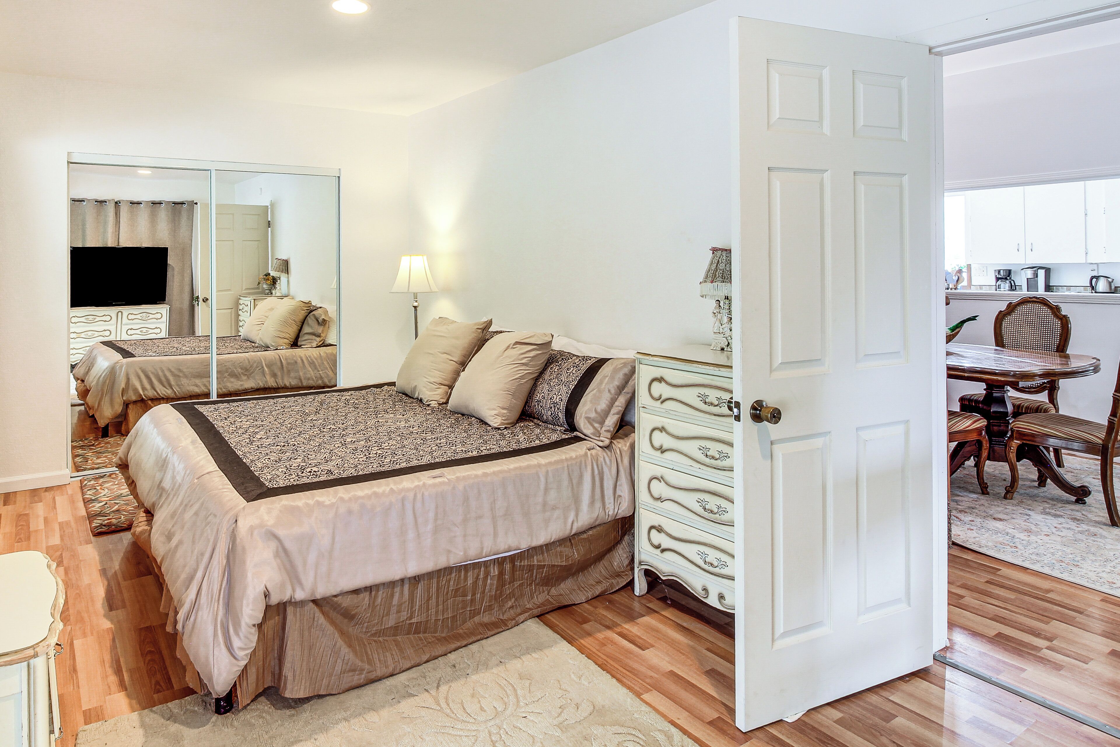 A bedroom with a bed, a dresser, a mirror, and a door leading to a dining area.