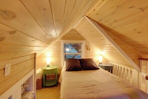 A cozy attic bedroom with a sloped ceiling, a bed with a beige comforter, two bedside lamps, and a window with a view of the outdoors. - Ski, Hike, Relax By Water: Rustic Mccall Tiny Home (McCall)