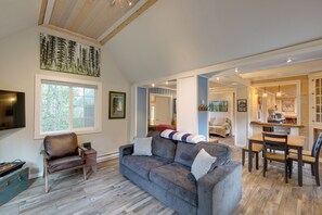 Ein geräumiger Wohnbereich mit grauem Sofa, Esstisch aus Holz und Stühlen. Ein gro ßes Fenster sorgt für natürliches Licht, und ein Fernseher ist an der Wand montiert.