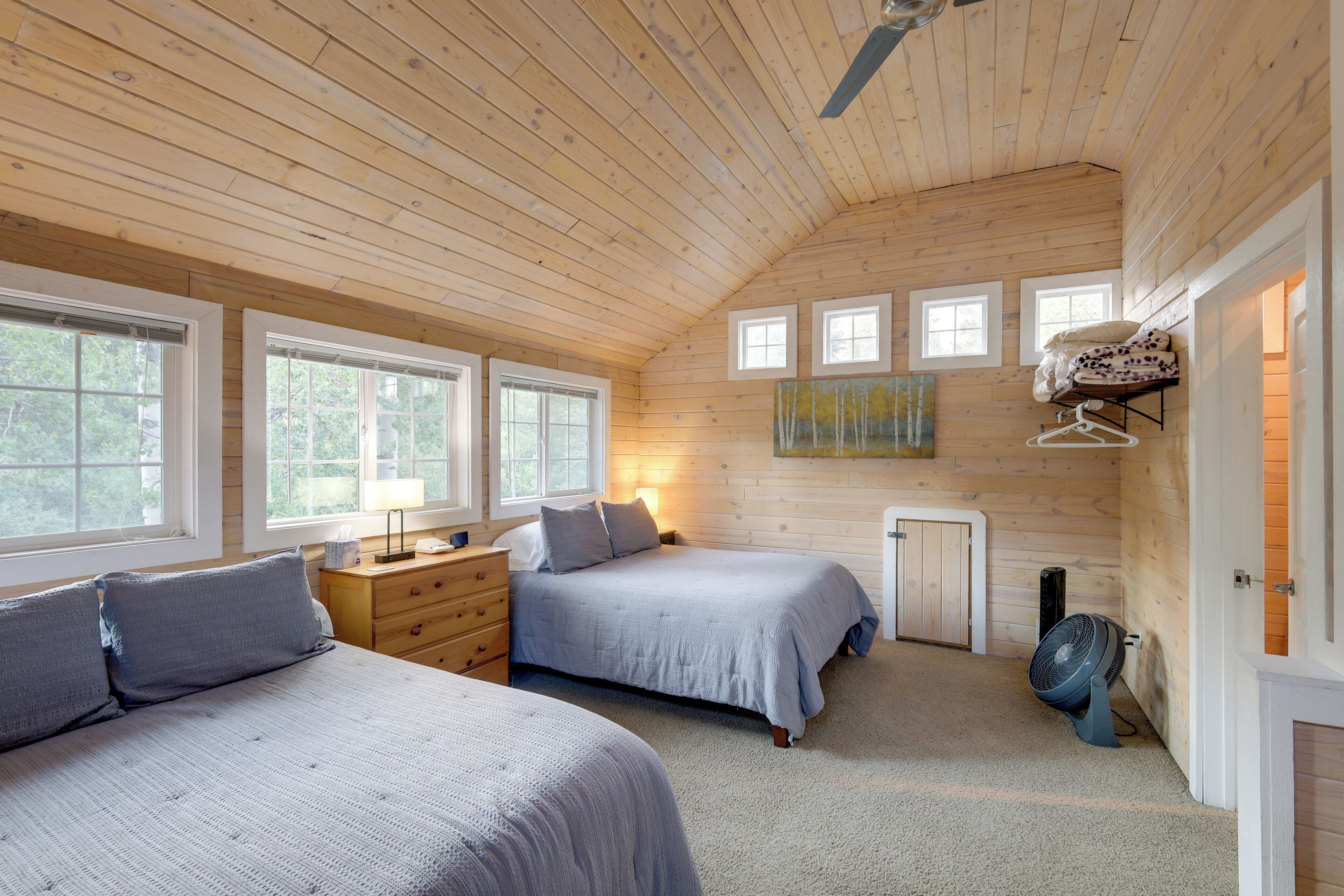 A cozy bedroom with two beds, a dresser, a fan, and a painting on the wall.