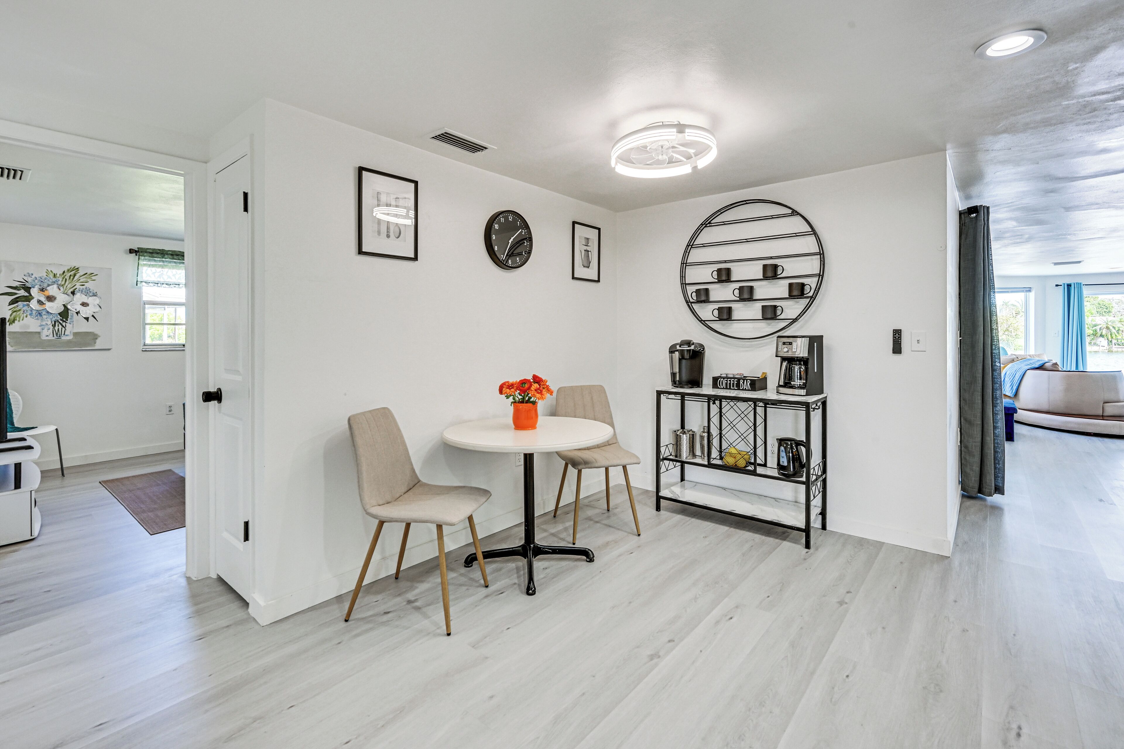 A modern, well-lit room with a round coffee table, two chairs, a coffee station, and a wall clock.