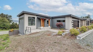A modern two-story house with a grey exterior, a wooden front door, and a small garden area with shrubs and a tree. - Fenced Yard, Water & Island Views: Anacortes Home! (Anacortes)