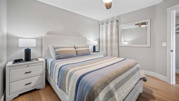 Une chambre à coucher comprenant un lit, des tables de chevet, un ventilateur de plafond, un miroir et une porte.