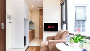 A modern living room with a sofa, a small table with a plant and water bottles, a TV displaying "NETFLIX", and a view of the outside through a window. - Studio in Modern building with AC; Kitchen; 1.3km from TSN airport (Hồ Chí Minh)