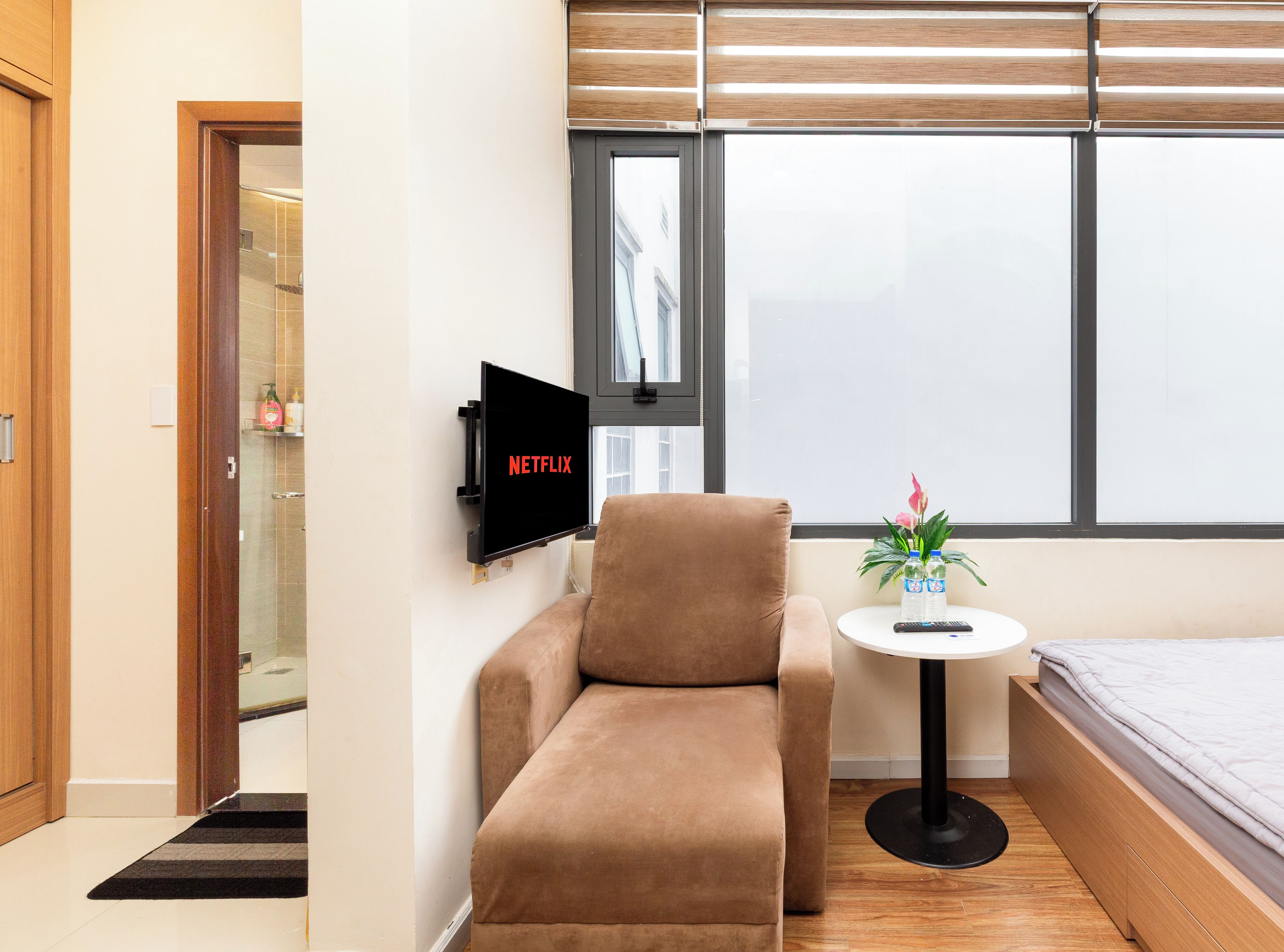Een moderne hotelkamer met een bank, een klein rond tafeltje met een afstandsbediening, een bed en een aan de muur bevestigde televisie.