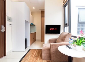 A modern living room with a sofa, a small table with a plant and water bottles, a TV displaying "NETFLIX", and a view of the outside through a window.