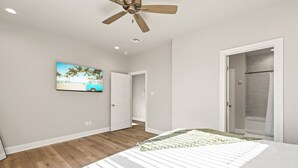 Un dormitorio con cama, ventilador de techo, un cuadro de una playa y una puerta que da a un baño.