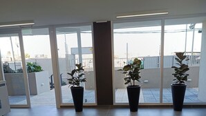 Une chambre d’hôtel moderne dotée de grandes portes-fenêtres coulissantes donnant sur un balcon, de plantes en pot, d’une petite table avec un ordinateur portable et d’une vue sur le paysage urbain.