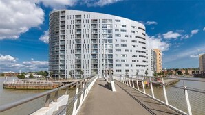 Ein modernes, mehrgeschossiges Gebäude mit einer geschwungenen Fassade, einer Fußgängerbrücke mit weißen Geländern und einem Fluss im Hintergrund.