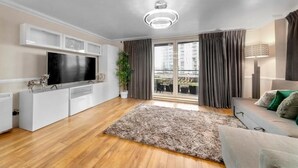 Ein modernes Wohnzimmer mit Flachbildfernseher, weißem Unterhaltungsschrank, grauem Sofa und einem großen Fenster mit Vorhängen.