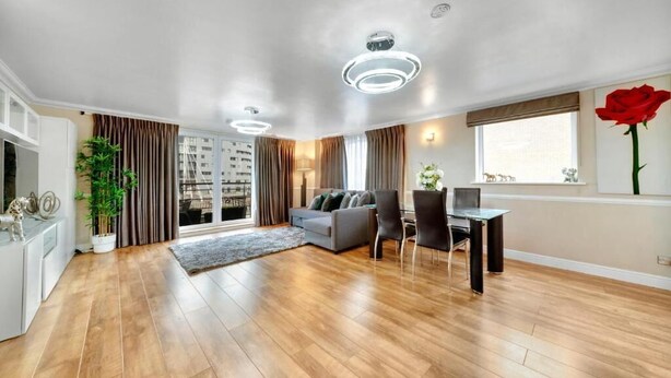 Ein modernes Wohnzimmer mit einem grauen Sofa, einem Esstisch mit Stühlen, einem großen Fenster mit Vorhängen und einer roten Rose auf der Fensterbank.