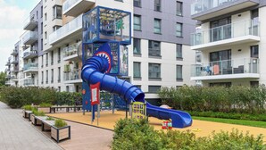Ein Spielplatz mit einer blauen Rutsche und Klettergerüsten, umgeben von einer angelegten Grünfläche mit Bänken und Pflanzen.