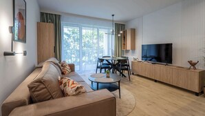 Un salon moderne avec un canapé, une table basse et un téléviseur. Il y a également un coin repas avec des chaises et une table. La pièce est dotée de grandes fenêtres et offre une vue sur les arbres.